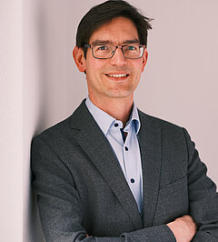 Portrait photo of Felix Höfling, smiling man with dark hair and glasses wearing a dark grey blazer and light blue shirt, arms crossed, light pink-white background.