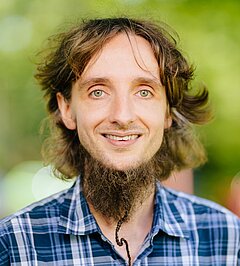 Portrait photo of Martin Brehm, smiling man with shoulder-length wavy brown hair and a braided beard wearing a blue checked shirt, blurred green outdoor background.