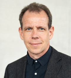 Portrait photo of Guido Schryen, man with short dark hair wearing a dark blazer and black shirt, light grey background.