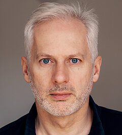 Portrait photo of Andreas Fink, man with short silver-white hair and light stubble wearing a dark navy shirt, neutral grey background.