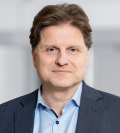 Portrait photo of Herbert Dawid, smiling man with dark brown hair wearing a dark navy blazer and light blue open-collar shirt, blurred light grey background.