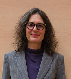 Portrait photo of Petra Imhof, smiling woman with long dark wavy hair and dark-framed glasses wearing a grey striped blazer and purple turtleneck, warm wooden wall background.