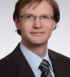 Portrait photo of Harald Köstler, smiling man with medium-length dark brown hair and thin-framed glasses wearing a dark brown blazer, light blue shirt and grey striped tie, light grey background.